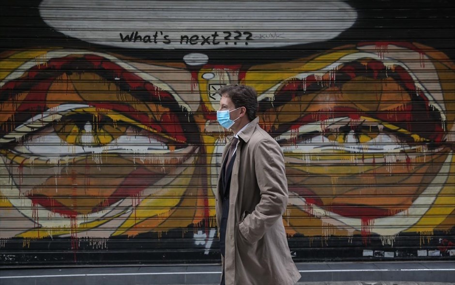 POY 2020. Ένας άντρας περπατάει στο κέντρο της Αθήνας φορώντας μάσκα. Πίσω του ένα γκράφιτι ρωτάει «Τι έπεται;»