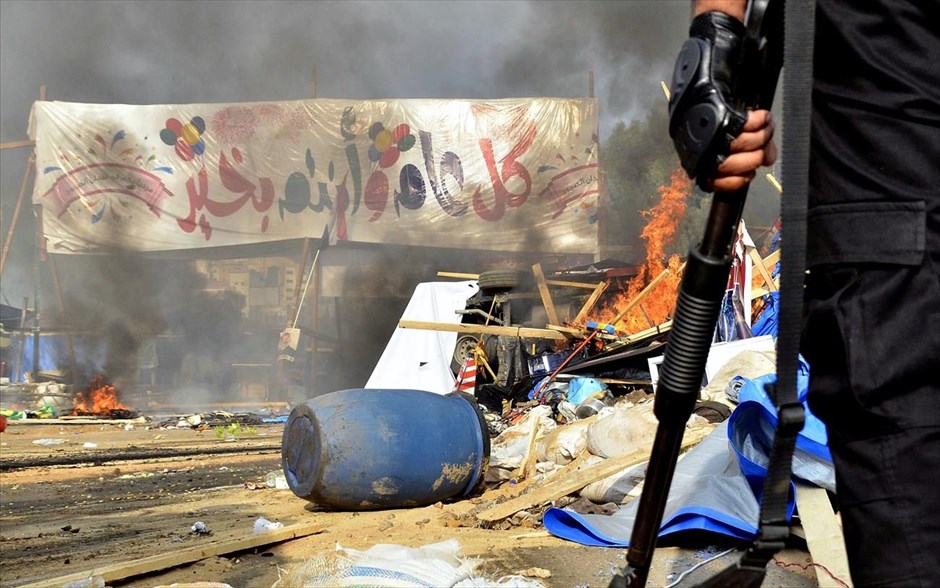 Αίγυπτος - Συγκρούσεις -  Ραμπάα αλ-Αλαουίγια. Πανό από τους πρόσφατους εορτασμούς για το Ιντ αλ-φιτρ διακρίνεται πίσω από τον καπνό και τα συντρίμμια κοντά στην πλατεία Ραμπάα αλ-Αλαουίγια, ύστερα από την επιχείρηση διάλυσης των συγκεντρώσεων υποστηρικτών του έκπτωτου προέδρου Μόρσι.