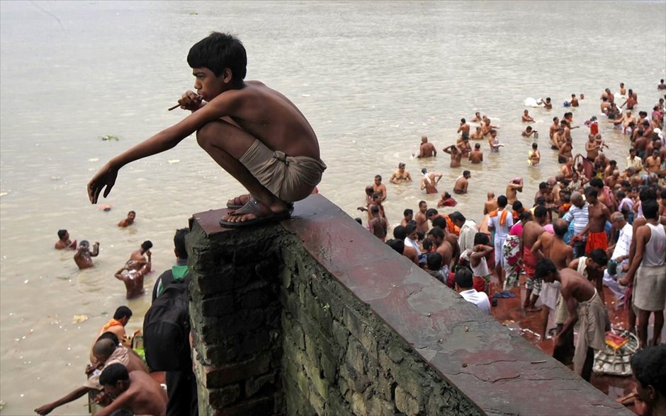 Ινδία: Προσευχές στον ποταμό Γάγγη. Ενας μικρός ινδός καθαρίζει τα δόντια του με ένα ξυλαράκι πάνω σε ένα τοίχο, παρατηρώντας τον κόσμο που βουτά στα νερά του ποταμού Γάγγη, την ιερή ημέρα Μαχαλάγια, στην Καλκούτα. Οι Ινδοί προσεύχονται αφού βουτήξουν στα νερά του ποταμού, τιμώντας τις ψυχές των προγόνων τους κατά τη διάρκεια της Μαχαλάγια, η οποία αποκαλείται επίσης Σράαντ ή Πίτρα Πάκσα.