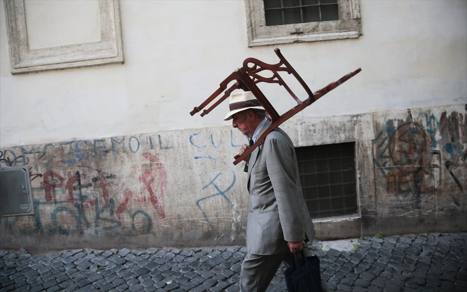 Στιγμιότυπο από τους δρόμους της Ρώμης. Ενας άντρας περπατά σε ένα δρόμο στη Ρώμη κρατώντας μία καρέκλα στους ώμους του.
