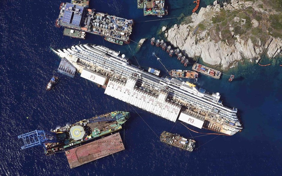 POY - Φωτογραφίες της χρονιάς - Αύγουστος 2013. Εμπορευματοκιβώτια έχουν τοποθετηθεί γύρω από το ναυάγιο του κρουαζιερόπλοιου Costa Concordia, στο πλαίσιο του σχεδίου ανέλκυσής του (26 Αυγούστου 2013). Το πλοίο ναυάγησε στις 13 Ιανουαρίου 2012 στα ανοιχτά του νησιού Τζίλιο, στην Τοσκάνη. Η προσπάθεια περιστροφής και ανόρθωσης του βυθισμένου κρουαζιερόπλοιου Costa Concordia ολοκληρώθηκε με επιτυχία στις 02.00 GMT στις 17 Σεπτεμβρίου έπειτα από 19 ώρες. Ήταν μια από τις πλέον πολύπλοκες και ακριβές επιχειρήσεις ανέλκυσης πλοίου που έγινε ποτέ καθώς το 114.500 τόνων κρουαζιερόπλοιο ανορθώθηκε με τη βοήθεια ειδικών χαλύβδινων καλωδίων και τοποθετήθηκε σε μια ειδική τεχνητή πλατφόρμα.