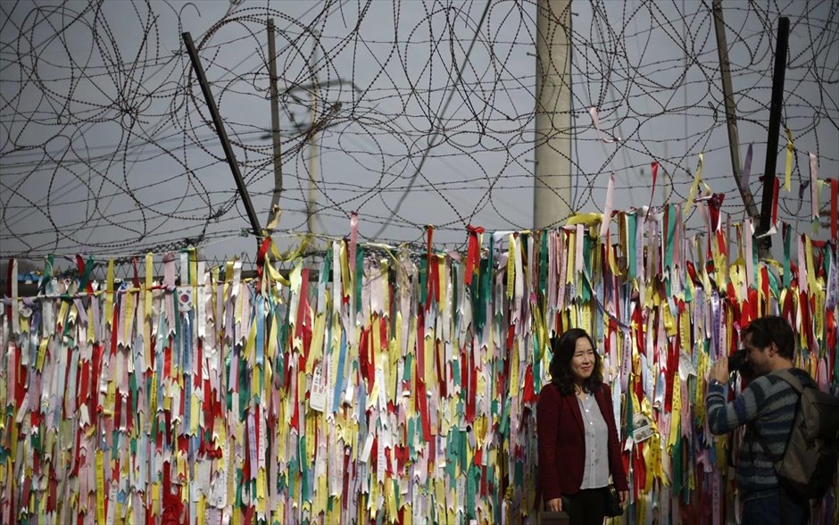 Τα τείχη που χωρίζουν τον κόσμο - Κορέα. Μία γυναίκα ποζάρει για μία αναμνηστική φωτογραφία μπροστά από το συρματόπλεγμα, διακοσμημένο με κορδέλες που φέρουν μηνύματα και ευχές για την ενοποίηση της Βόρειας και της Νότιας Κορέας, κοντά στην αποστρατικοποιημένη ζώνη στο Πατζού. Η αποστρατικοποιημένη ζώνη καθορίστηκε το 1953, όταν υπογράφηκε ανακωχή μεταξύ Βόρειας και Νότιας Κορέας. Έχει μήκος 248 χιλιόμετρα και αποτελείται από ηλεκτροφόρους φράκτες. Φυλάσσεται από 700.000 στρατιώτες από τη Βόρεια Κορέα και περισσότερους από 400.000 από τη Νότια Κορέα.