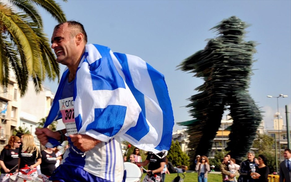 32ος Μαραθώνιος Αθήνας. Αθλητής κρατώντας την ελληνική σημαία περνά μπροστά από το άγαλμα του Δρομέα.