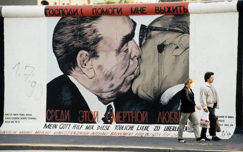 Η πτώση του Τείχους του Βερολίνου. Ένα ζευγάρι περνά μπροστά μία τοιχογραφία στο Τείχος του Βερολίνου, του Ρώσου καλλιτέχνη Dmitrij Vrubel (φωτογραφία αρχείου, Ιούνιος 1990). Η τοιχογραφία απεικονίζει ένα φιλί μεταξύ του Γενικού Γραμματέα του Κομμουνιστικού Κόμματος της ΕΣΣΔ Λεονίντ Μπρέζνιεφ και του ομολόγου του Έριχ Χόνεκερ (δεξιά) από τη Λαϊκή Δημοκρατία της Γερμανίας.