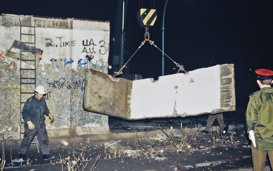 Η πτώση του Τείχους του Βερολίνου. Αξιωματικός της στρατιωτικής αστυνομίας των Συμμαχικών Δυνάμεων επιβλέπει καθώς συνοριακά στρατεύματα της Ανατολικής Γερμανίας διαλύουν με βαριά μηχανήματα τμήματα του τείχους του Βερολίνου και καθαρίζουν τα μπάζα από την ανατολική γερμανική πλευρά του Τείχους, κοντά στην Πύλη του Βραδεμβούργου (Φωτογραφία αρχείου, Ιανουάριος 1990).