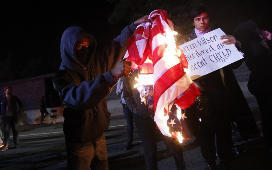 Φέργκιουσον - Επεισόδια μετά την απόφαση για τον αστυνομικό - Καλιφόρνια. Διαδηλωτής καίει μία αμερικανική σημαία στον αυτοκινητόδρομο 580, στο Όκλαντ της Καλιφόρνια, κατά τη διάρκεια διαδήλωσης μετά την ανακοίνωση της απόφασης του σώματος των ενόρκων να μην παραπεμφθεί σε δίκη ο αστυνομικός Ντάρεν Ουίλσον, που στις 9 Αυγούστου σκότωσε τον άοπλο 18χρονο Αφροαμερικανό Μάικλ Μπράουν.