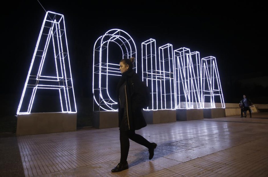 Χριστουγεννιάτικες βόλτες στο κέντρο της Αθήνας. Μία γυναίκα περπατάει μπροστά από το φωτισμένο σήμα της Αθήνας, μέρος του χριστουγεννιάτικου στολισμού της πόλης.