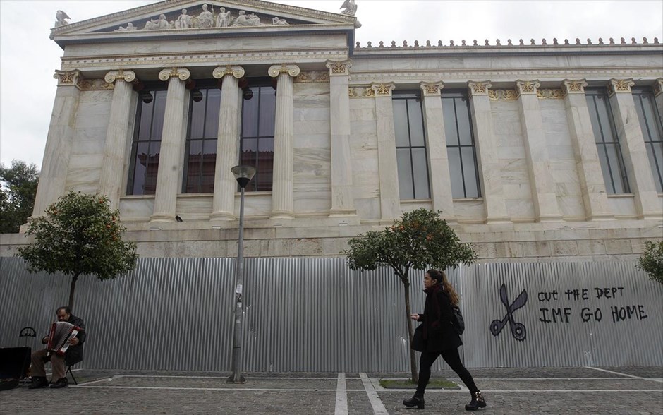 Στιγμιότυπο από την Αθήνα της κρίσης . Πλανόδιος μουσικός παίζει το ακορντεόν του μπροστά από τοίχο στον οποίο έχει γραφτεί με μπογιά (στα αγγλικά) η φράση: «Κόψτε το χρέος - ΔΝΤ φύγε».