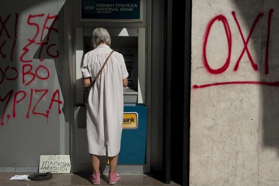 Στον ρυθμό του δημοψηφίσματος - Τράπεζες - ATM. Μια γυναίκα κάνει ανάληψη από ATM της Εθνικής Τράπεζας. Στον διπλανό τοίχο υπάρχει γκράφιτι υπέρ του «Όχι», ενώ από την άλλη πλευρά διακρίνονται τα αντικείμενα ενός επαίτη.